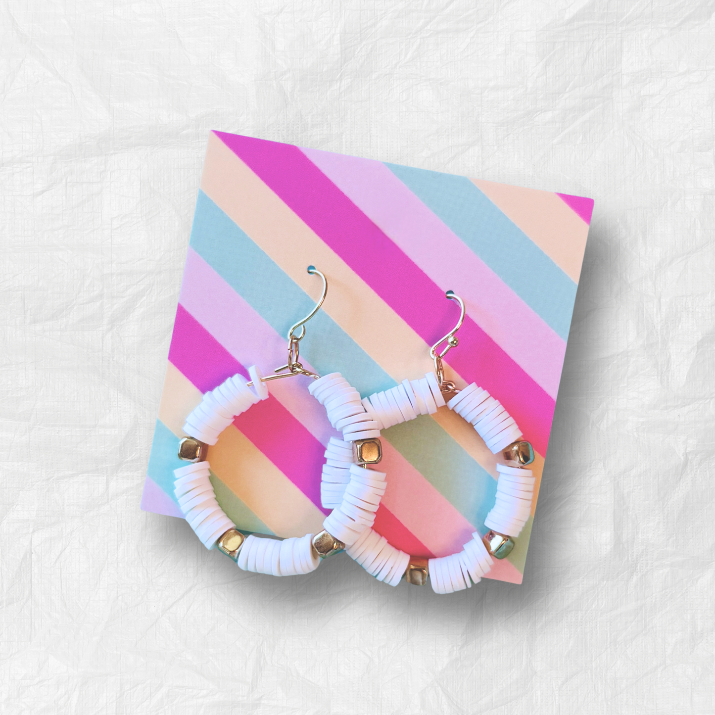 A pair of white and gold hoop earrings with heishi beads, inspired by coastal elegance, displayed against a soft blue gradient background for a minimal yet luxurious look.
