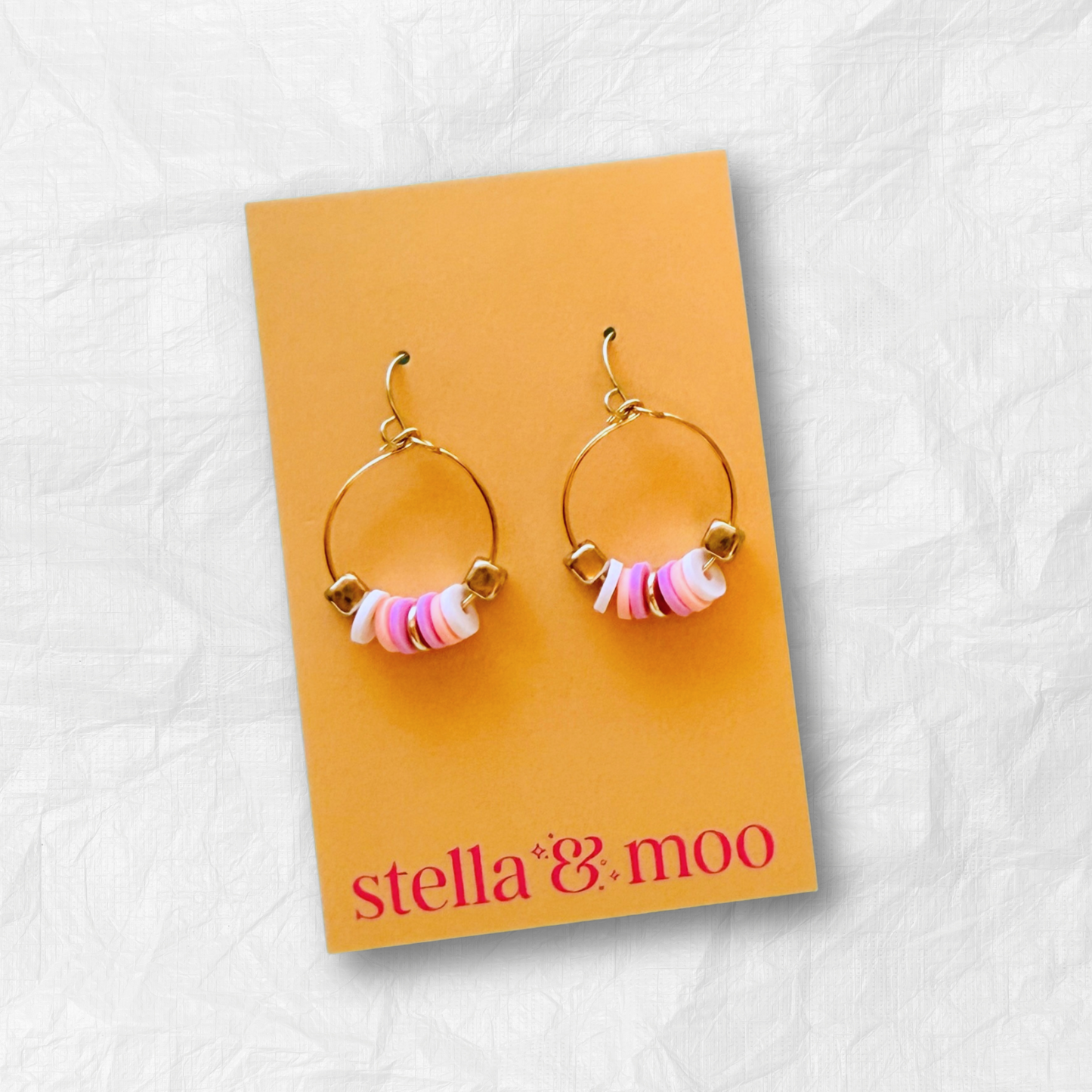 A pair of delicate gold mini hoop earrings featuring soft pink and white beaded accents, presented on a warm-toned Stella & Moo branded card, perfect for a fun yet minimal statement look.