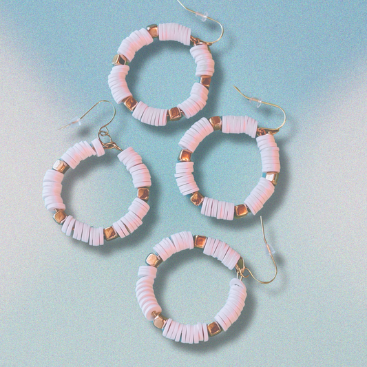 A pair of white and gold hoop earrings with heishi beads, inspired by coastal elegance, displayed against a soft blue gradient background for a minimal yet luxurious look.