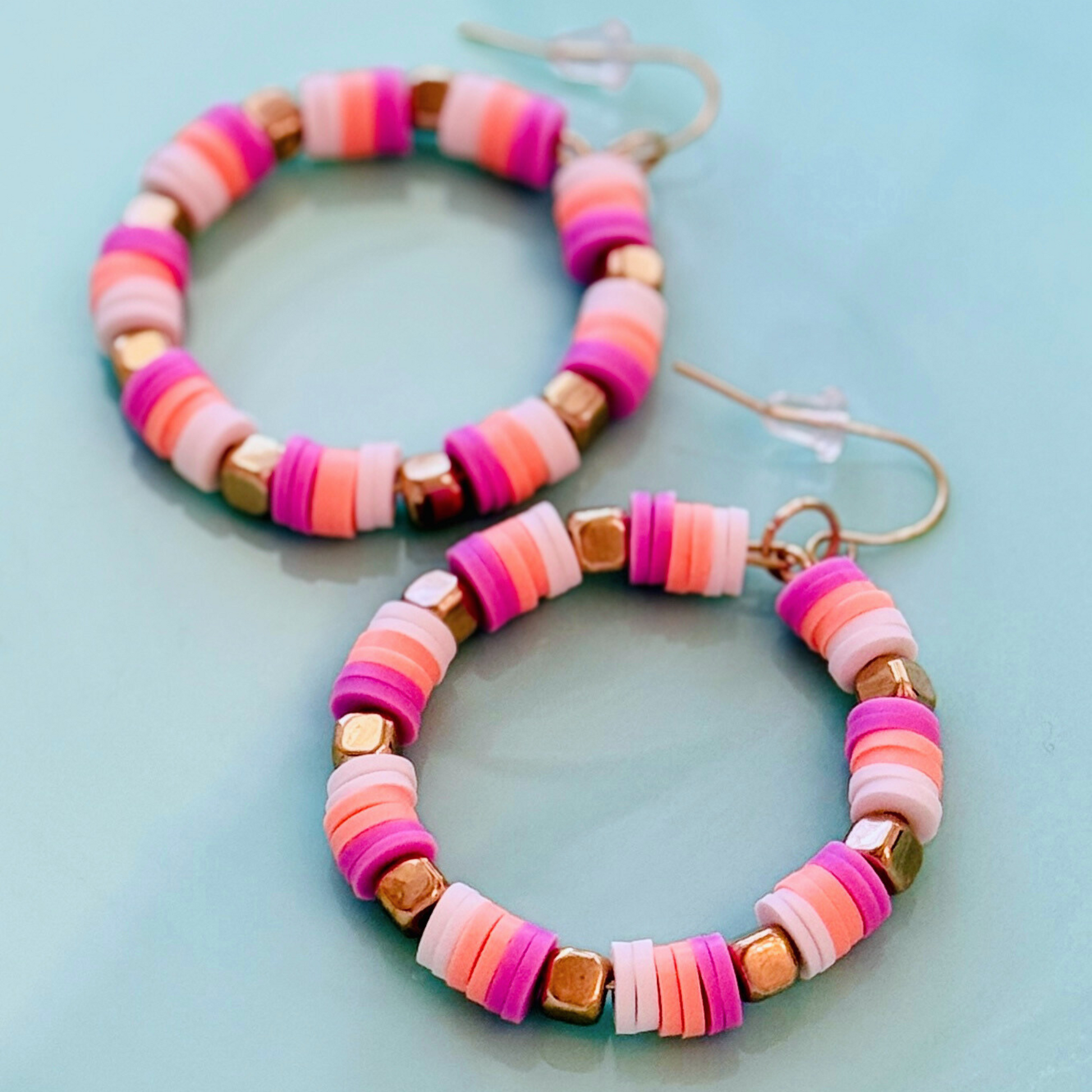 A close-up of handcrafted hoop earrings featuring soft pruple, creamsickle orange, and white beads with gold accents, inspired by the colors of French macarons, displayed on a soft blue background for a fun and playful statement look.