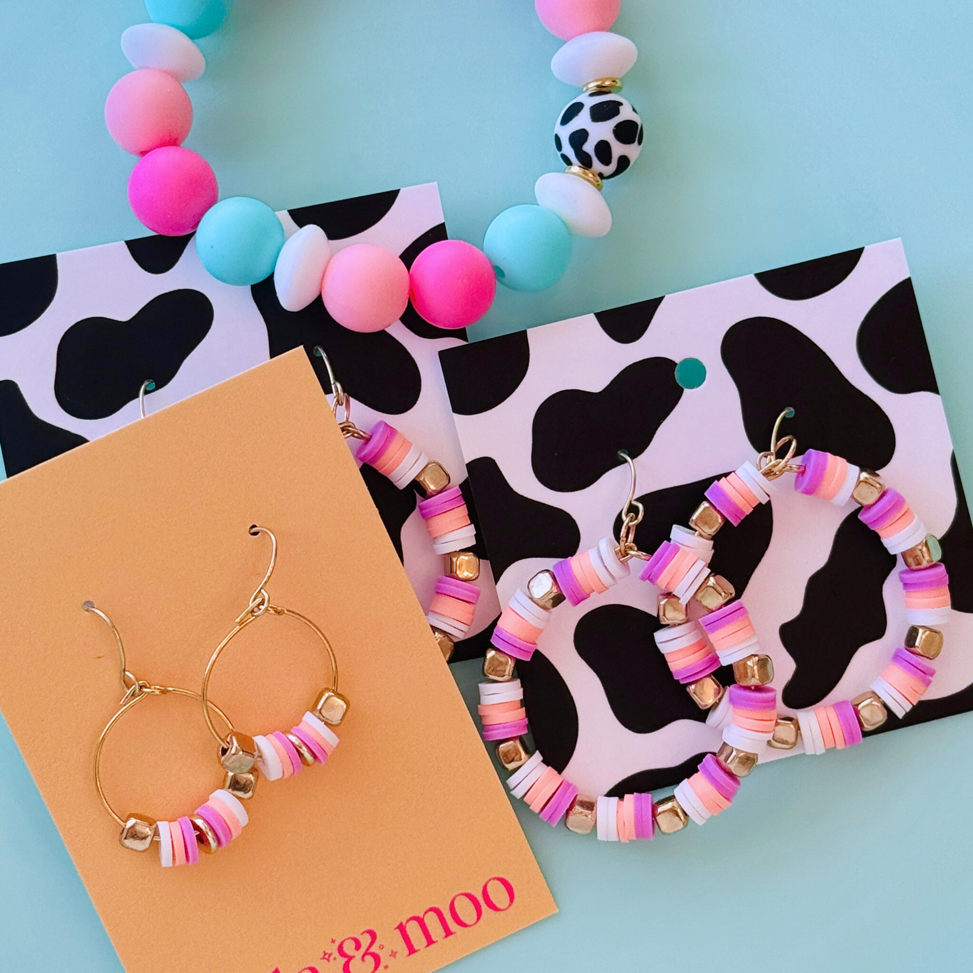 A close-up of handcrafted hoop earrings featuring soft pruple, creamsickle orange, and white beads with gold accents, inspired by the colors of French macarons, displayed on a soft blue background for a fun and playful statement look.